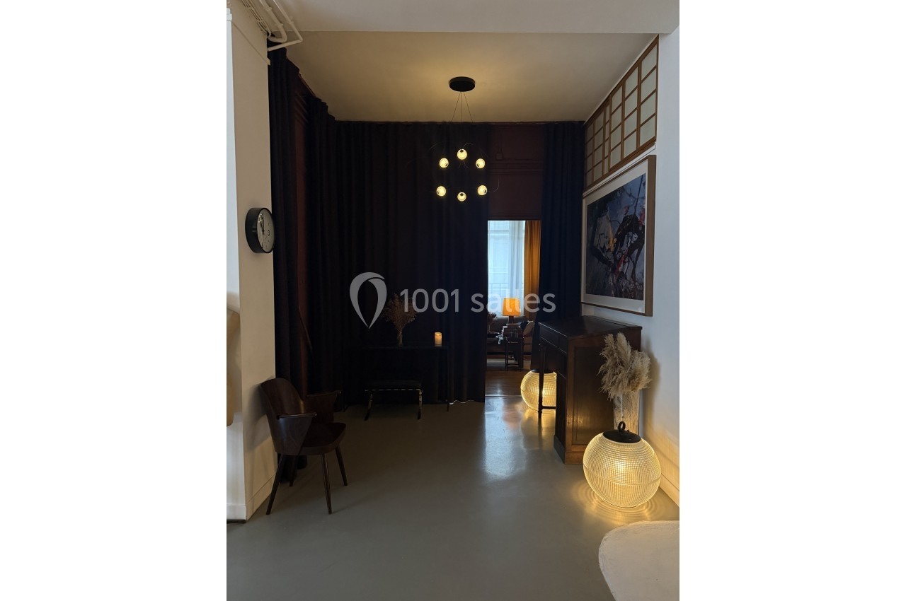 Intérieur sombre avec rideaux noirs, chaises en bois, luminaires suspendus et lampes au sol diffusant une lumière douce.