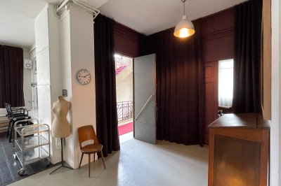 Une chaise en bois et un mannequin près d'une porte ouverte donnant sur un couloir lumineux avec une rambarde en fer forgé.