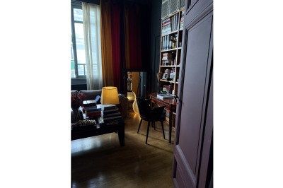 Une chaise en bois et un mannequin près d'une porte ouverte donnant sur un couloir lumineux avec une rambarde en fer forgé.