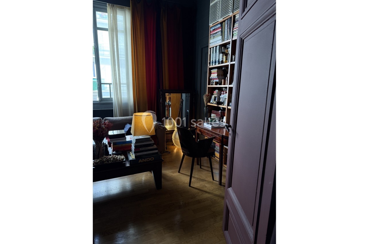 Pièce sombre avec bibliothèque, bureau en bois, lampe allumée et fauteuil, près d'une fenêtre avec rideaux.