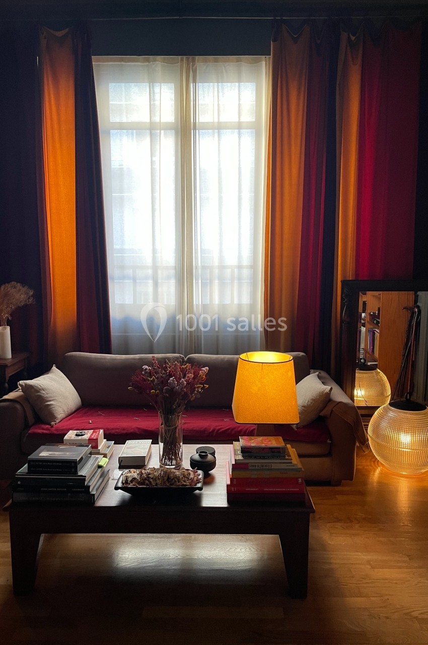 Salon avec canapé beige, table basse chargée de livres, lampe allumée et rideaux colorés devant une fenêtre.