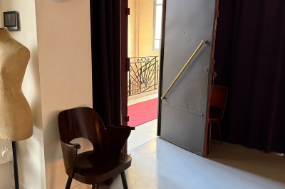 Une chaise en bois et un mannequin près d'une porte ouverte donnant sur un couloir lumineux avec une rambarde en fer forgé.