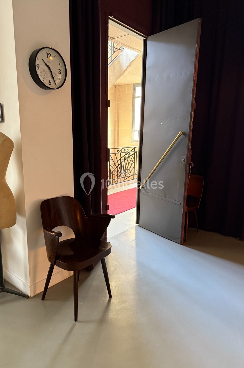Une chaise en bois et un mannequin près d'une porte ouverte donnant sur un couloir lumineux avec une rambarde en fer forgé.