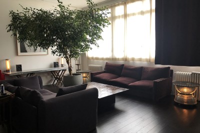Une chaise en bois et un mannequin près d'une porte ouverte donnant sur un couloir lumineux avec une rambarde en fer forgé.