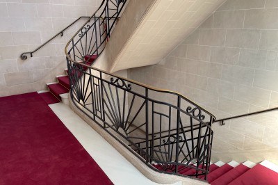Escalier intérieur en pierre avec rampe en fer forgé, tapis rouge et grande fenêtre laissant entrer la lumière naturelle.