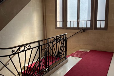 Escalier intérieur en pierre avec rampe en fer forgé, tapis rouge et grande fenêtre laissant entrer la lumière naturelle.