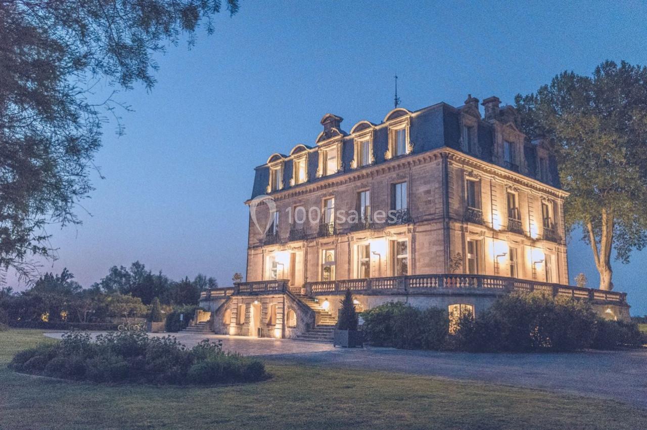 Manoir éclairé au crépuscule, entouré de verdure et d'arbres, avec un ciel bleu en arrière-plan.