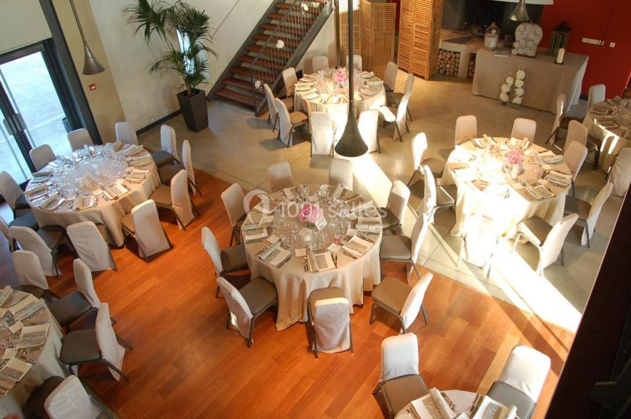 Salle de réception lumineuse avec tables rondes dressées, nappes beige et décorations florales roses.