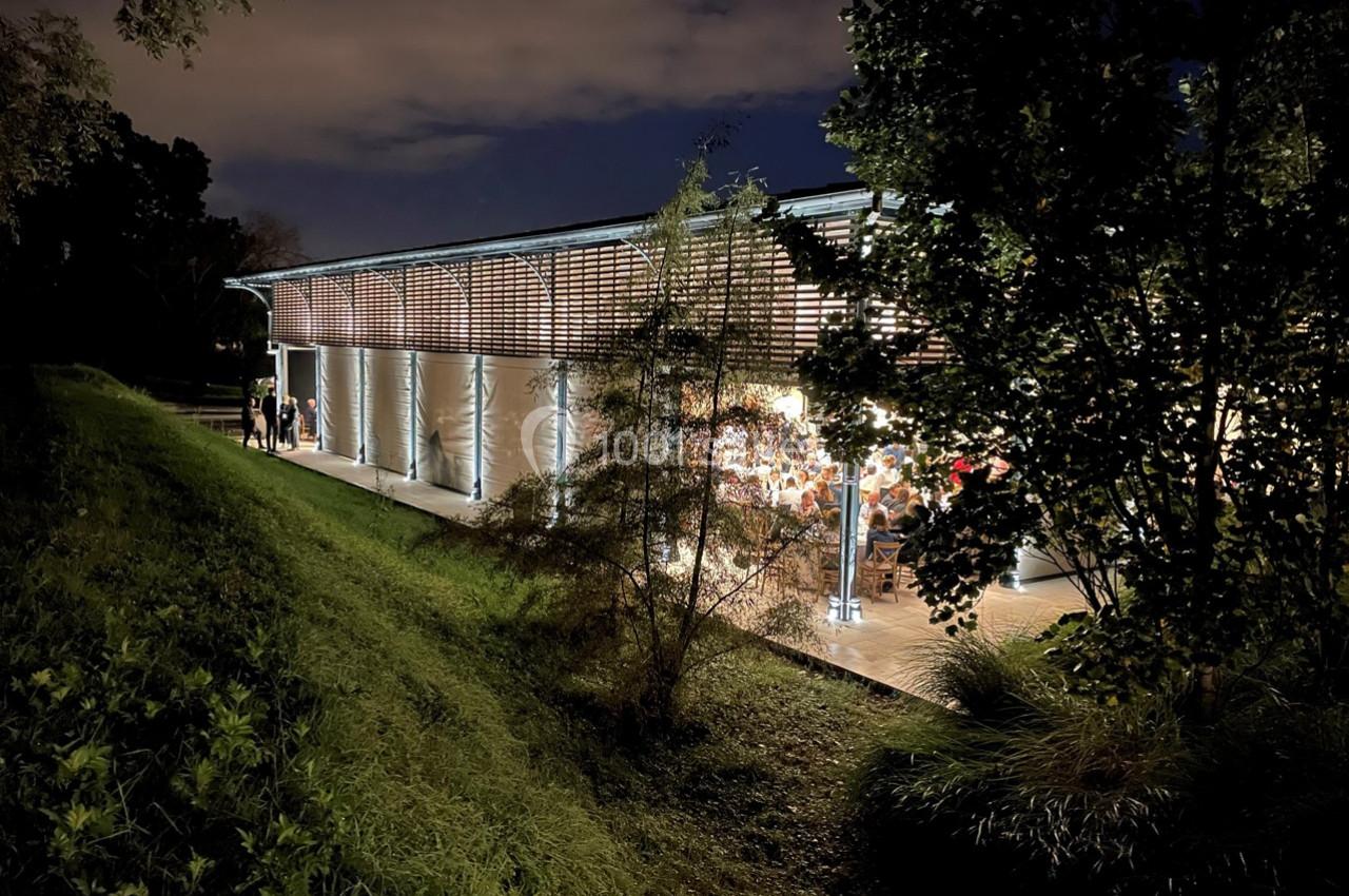 Bâtiment moderne éclairé de nuit, entouré d'arbres et de végétation, avec des personnes visibles à l'intérieur.