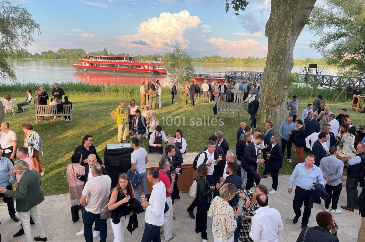 Groupe de personnes rassemblées en extérieur près d'un fleuve, avec un bateau rouge amarré en arrière-plan.