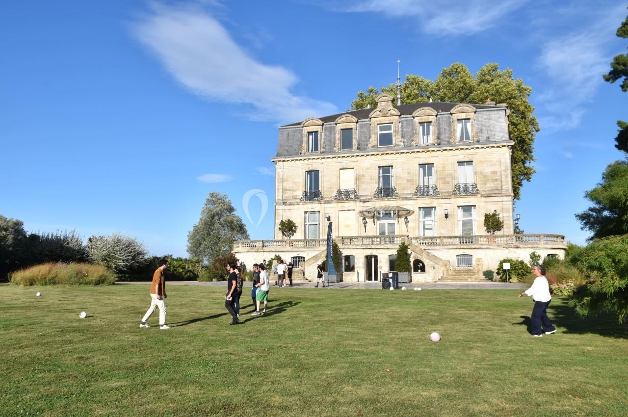Un groupe de personnes joue au ballon sur une pelouse devant un grand bâtiment ancien sous un ciel dégagé.