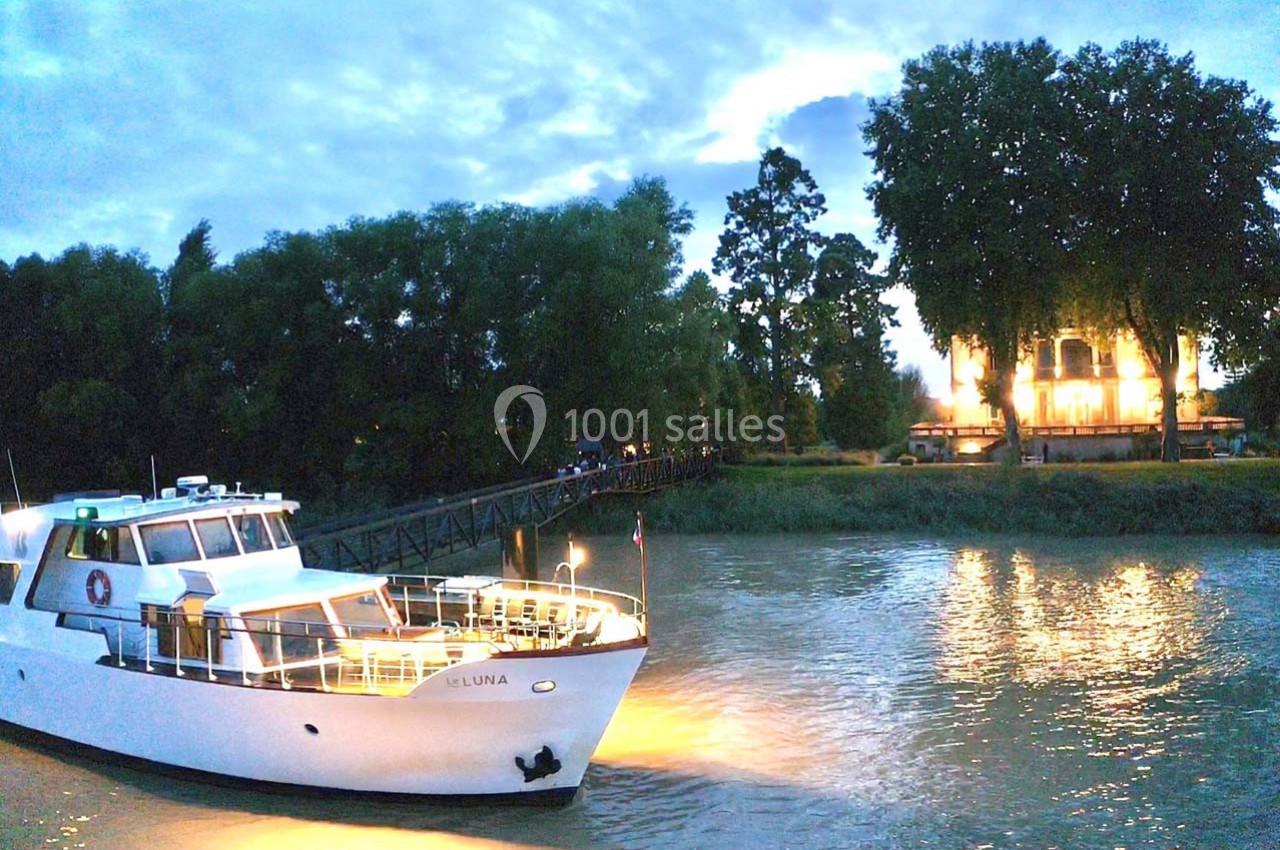 Bateau amarré sur une rivière au crépuscule, avec un bâtiment éclairé et des arbres en arrière-plan.