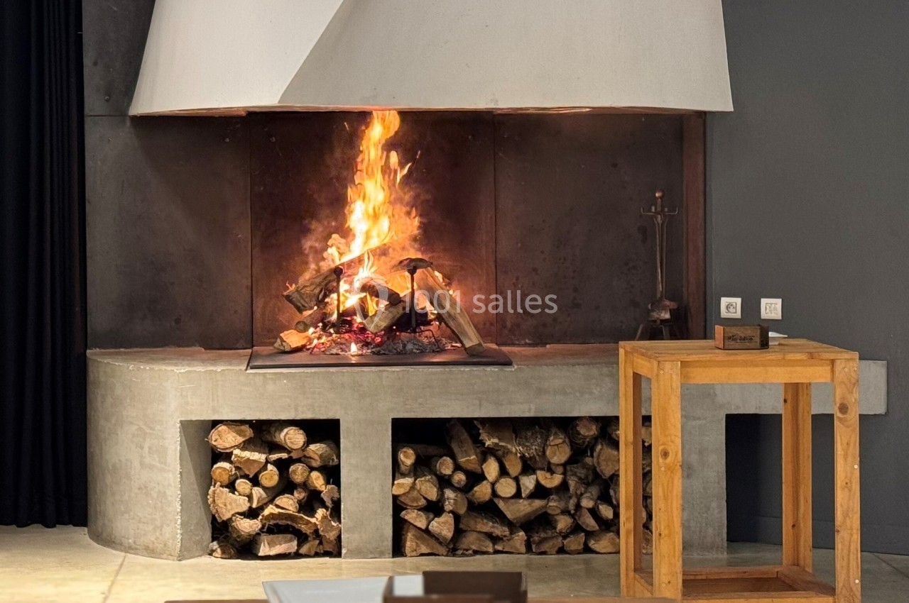 Cheminée moderne allumée avec bûches empilées en dessous, entourée de mobilier en bois dans une pièce sobre.