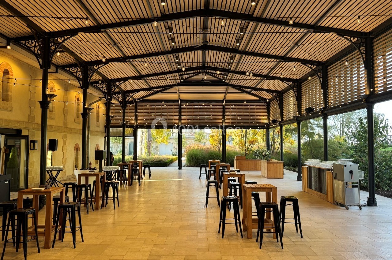 Salle lumineuse avec toit en verre, tables hautes en bois et tabourets noirs, entourée de verdure extérieure.