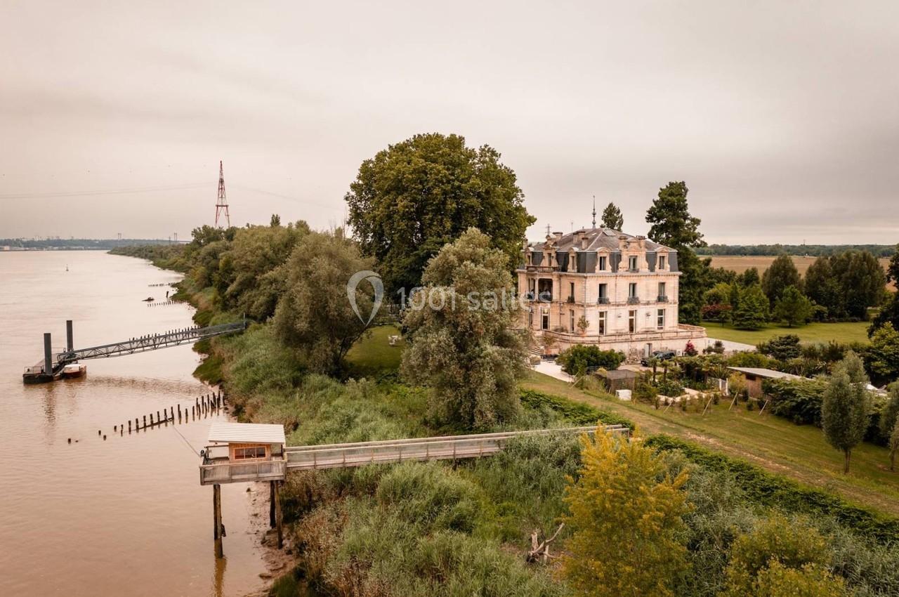 Vue aérienne d'une maison ancienne entourée de verdure, située près d'un fleuve avec un ponton en bois.