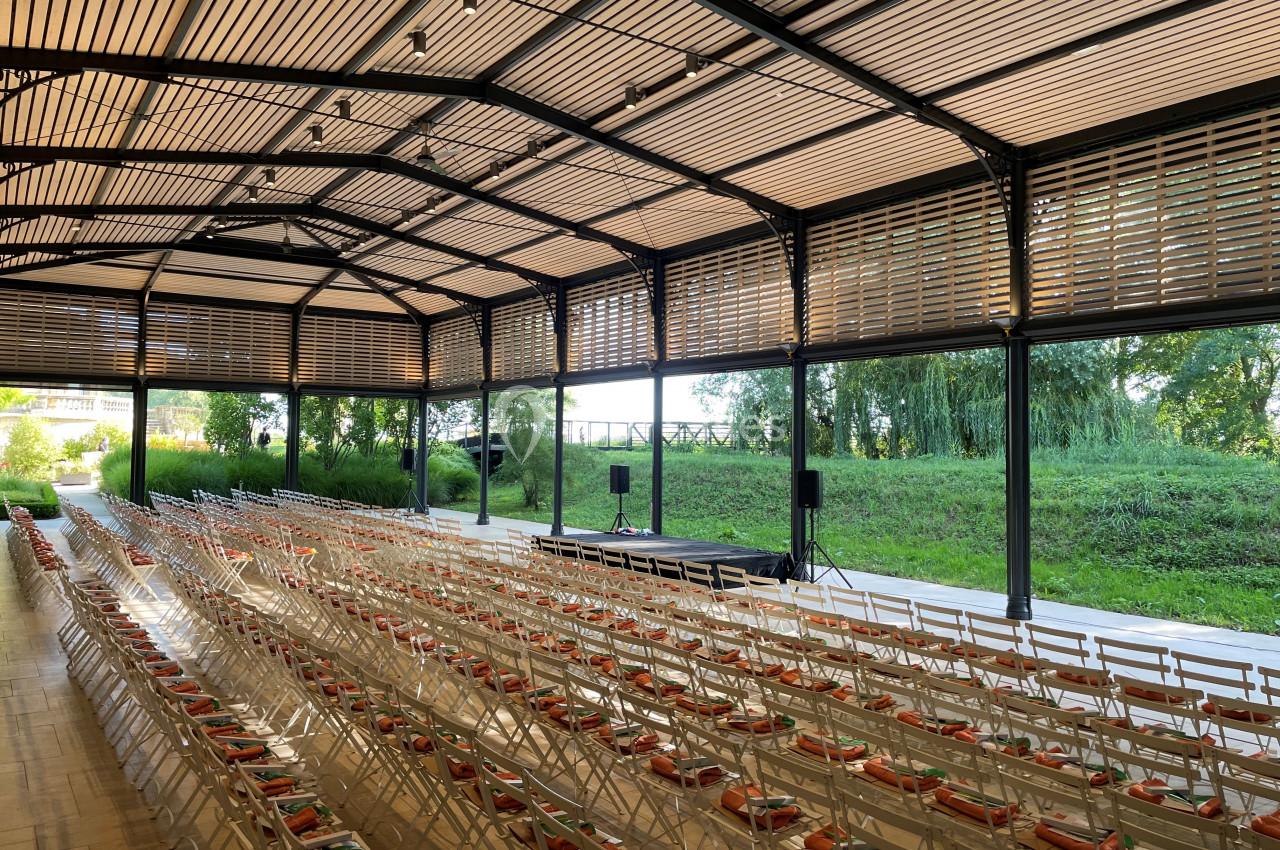 Salle extérieure couverte avec rangées de chaises pliantes face à une petite scène, entourée de verdure.