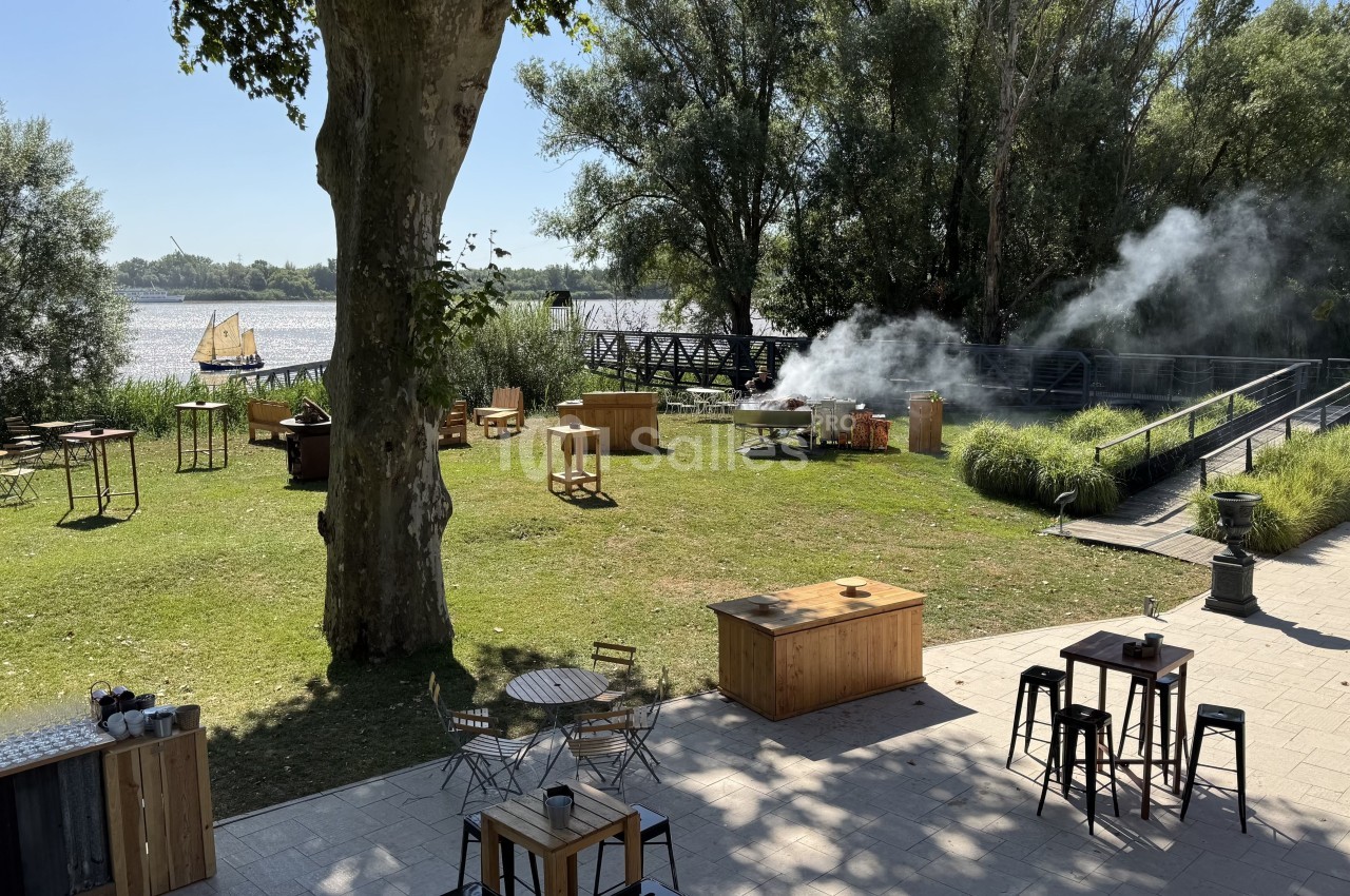 Espace extérieur verdoyant avec tables, chaises et fumée près d'un barbecue, vue sur un fleuve et un voilier au loin.