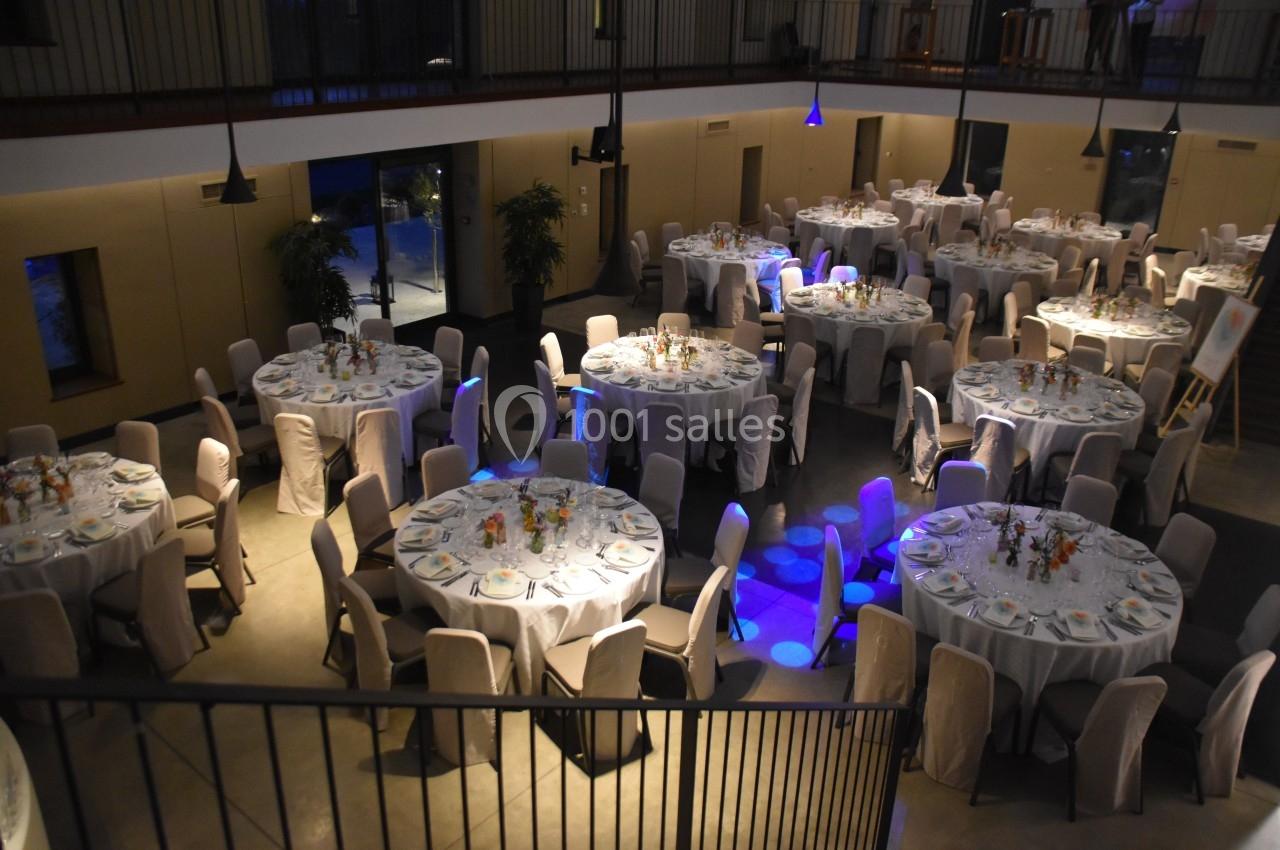Salle de réception avec tables rondes dressées, nappes blanches et chaises couvertes, éclairage tamisé.