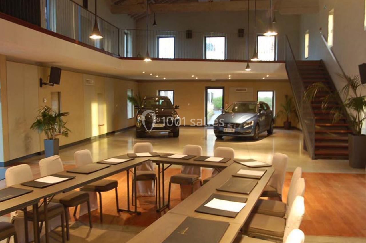 Salle de réunion lumineuse avec une grande table, des chaises et deux voitures exposées en arrière-plan.