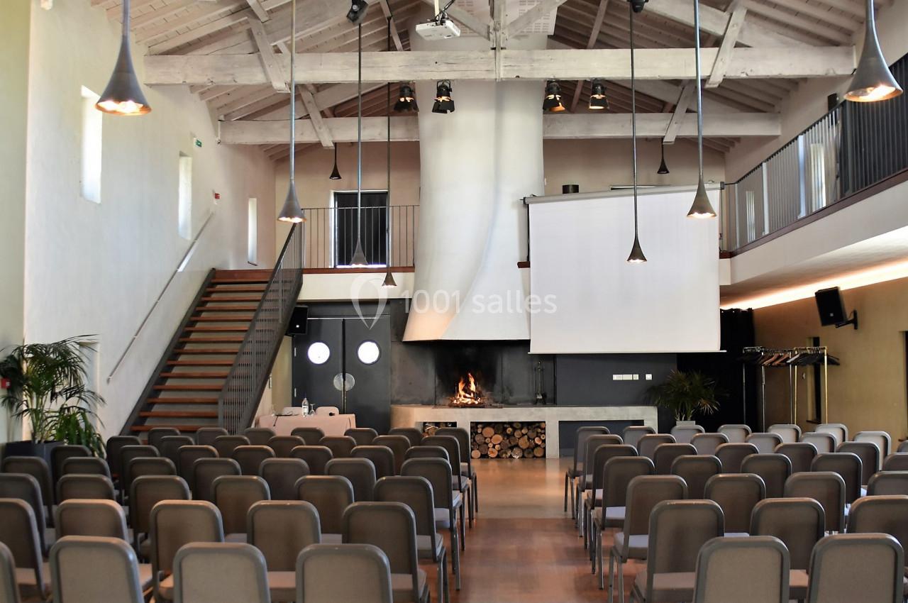 Salle de conférence lumineuse avec rangées de chaises, écran de projection, cheminée et mezzanine.