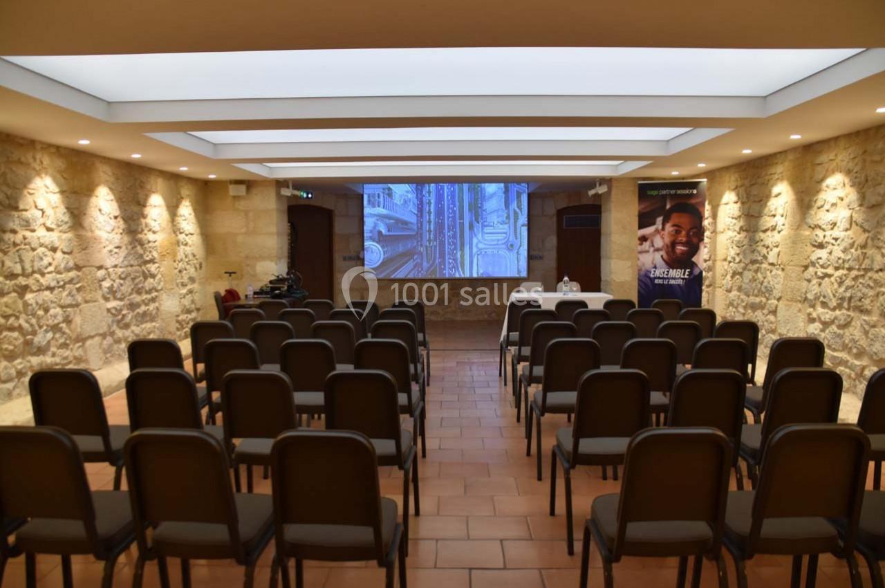 Salle de conférence avec des rangées de chaises noires, un écran de projection et des murs en pierre apparente.