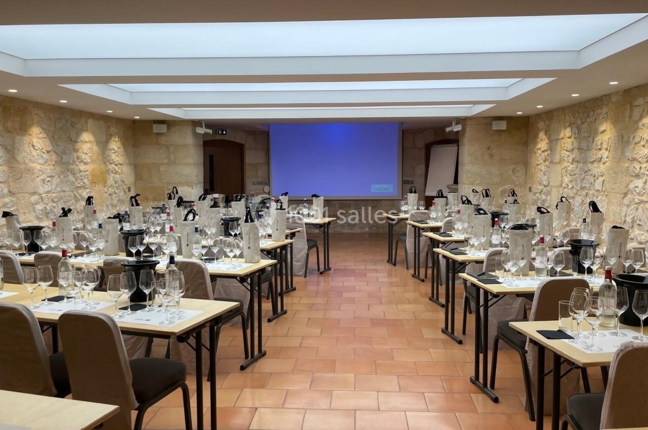 Salle de dégustation aménagée avec des tables, des verres, des bouteilles et un écran de projection au fond.