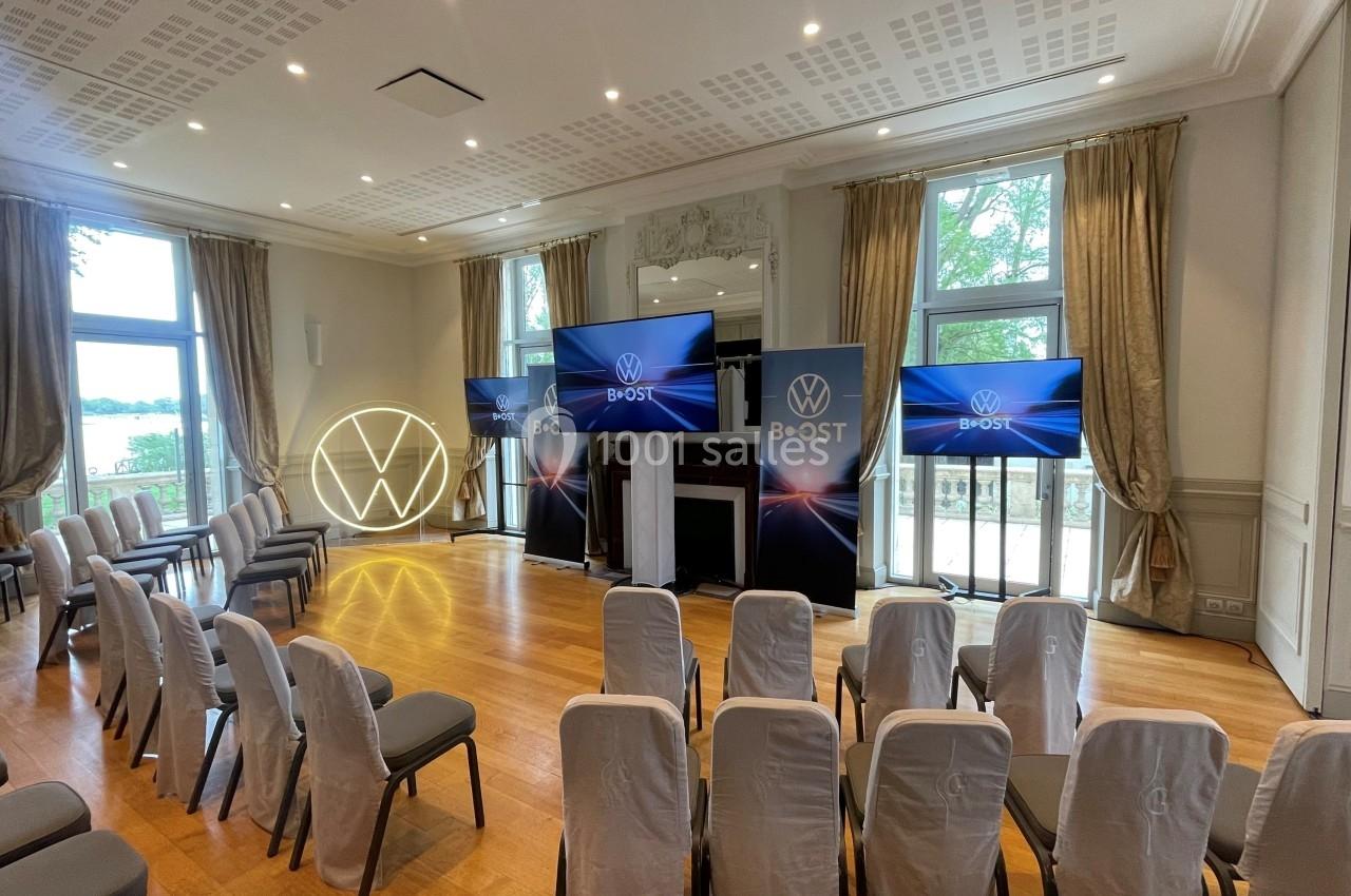 Salle de conférence lumineuse avec chaises alignées, écrans et logo lumineux VW, donnant sur des fenêtres ouvertes.