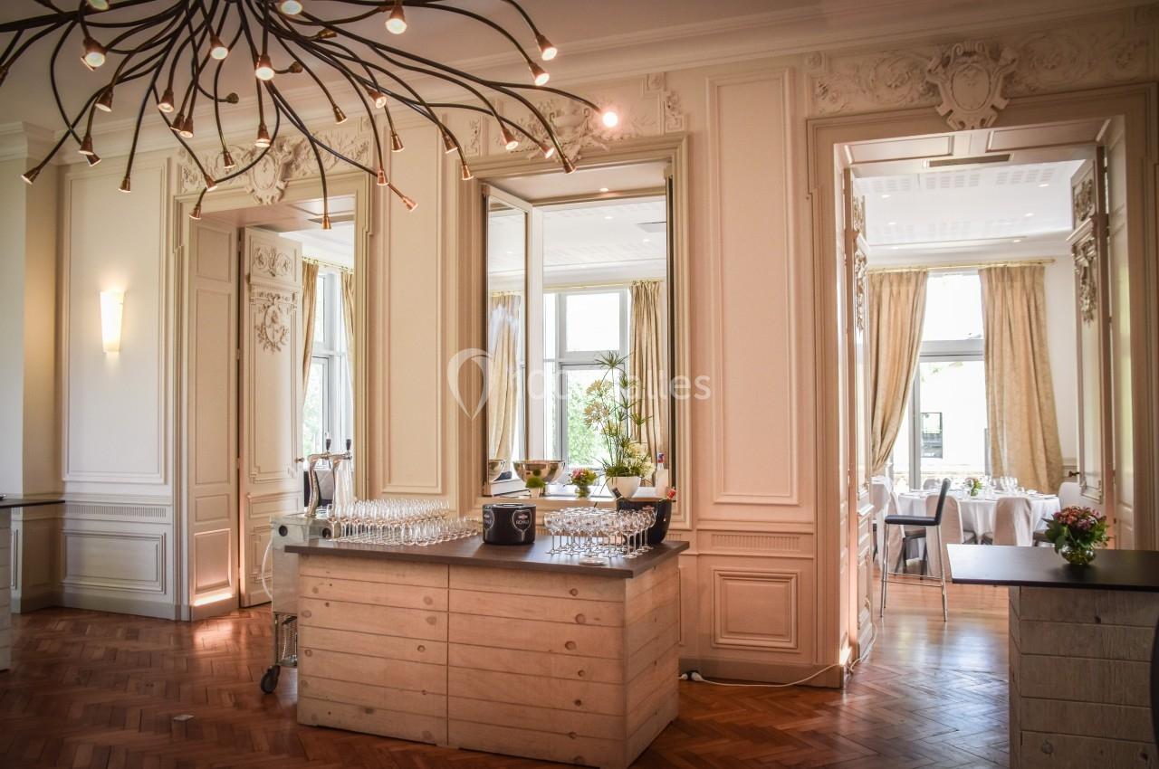 Salle lumineuse avec parquet, moulures, grandes fenêtres et un comptoir avec des verres et des bouteilles.