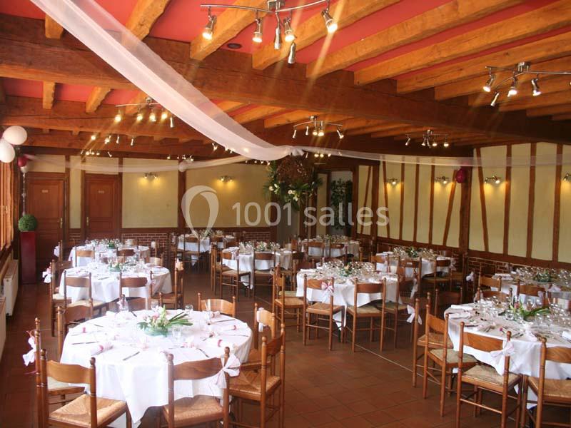 Salle de réception décorée avec des tables rondes dressées, chaises en bois et poutres apparentes au plafond.
