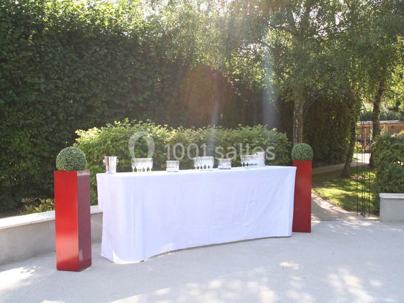 Table drapée de blanc avec des verres et des seaux à glace, entourée de verdure et de deux colonnes rouges décoratives.