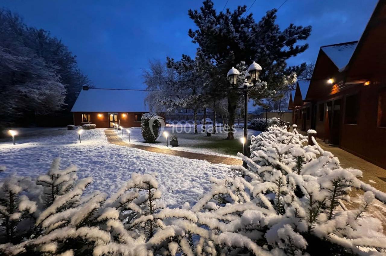 Allée bordée de buissons enneigés, éclairée par des lampadaires, avec des chalets en bois sous un ciel nocturne.