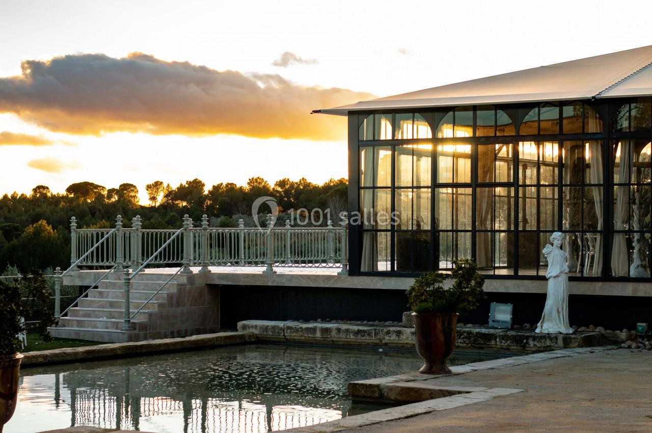 Verrière élégante au bord d'un bassin, entourée de végétation, avec un coucher de soleil en arrière-plan.