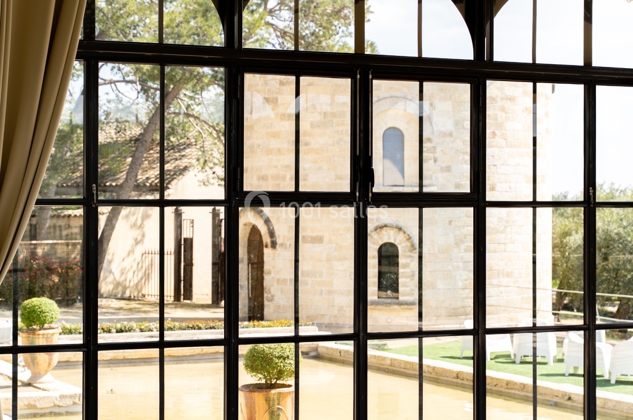 Vue sur une cour avec bassin et bâtiment en pierre, capturée à travers une grande baie vitrée en métal noir.