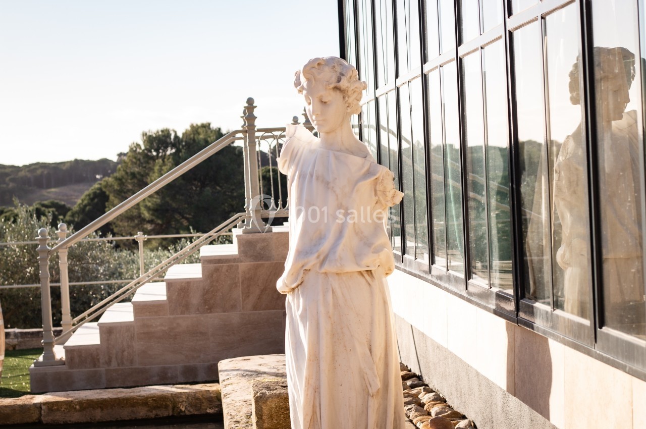 Statue en marbre représentant une femme, placée près d'une verrière avec un escalier et un paysage naturel en arrière-plan.