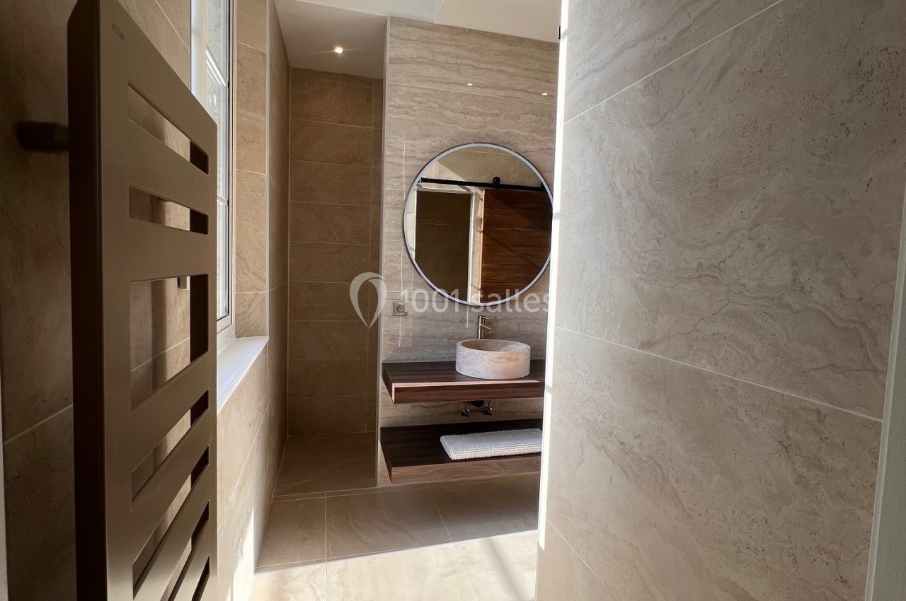 Salle de bain lumineuse avec murs en carrelage beige, miroir rond, vasque sur meuble en bois et sèche-serviettes.