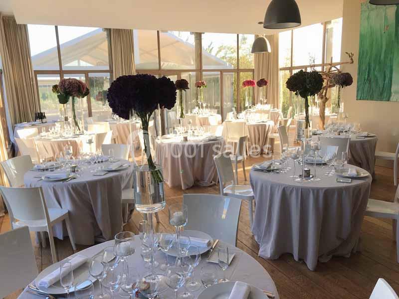 Salle de réception lumineuse avec tables rondes dressées, nappes grises et centres de table fleuris.