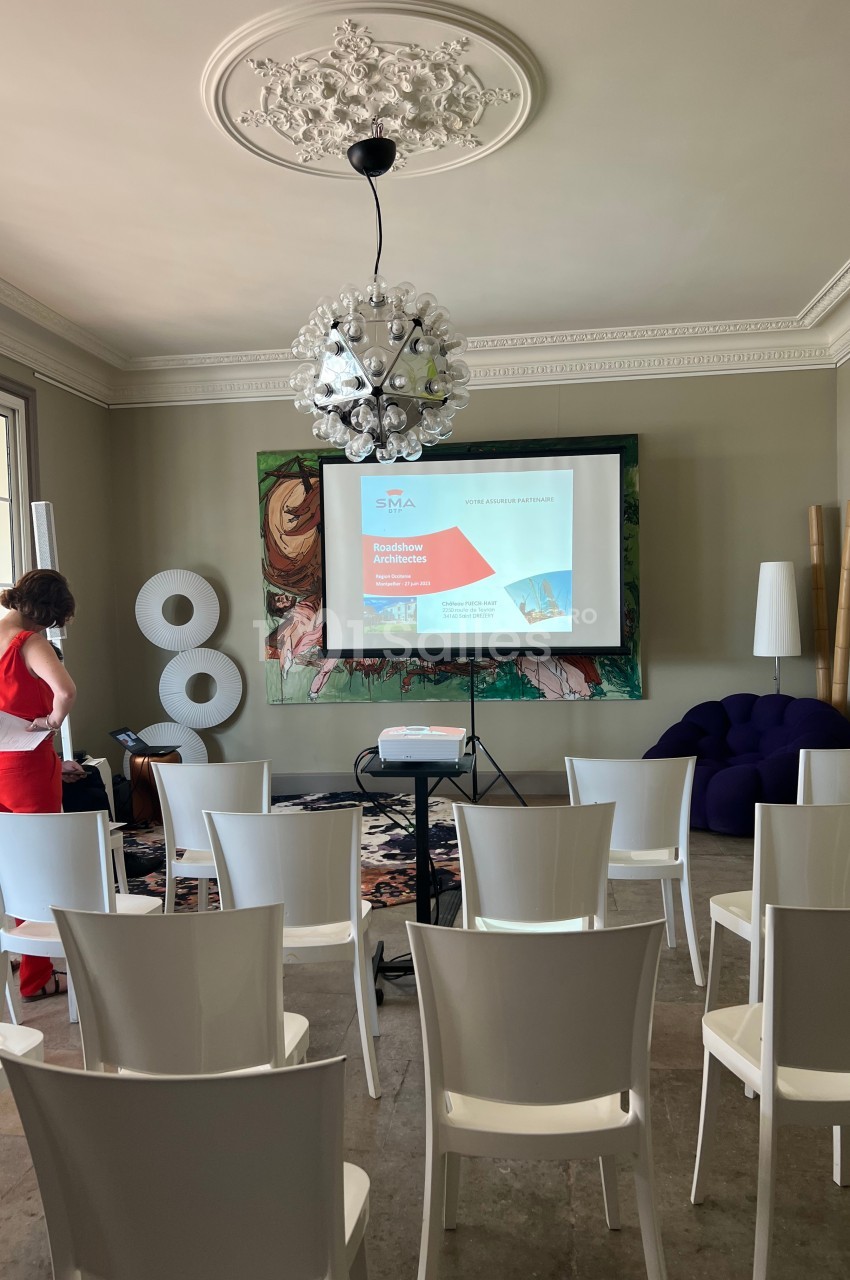 Salle de conférence lumineuse avec des chaises blanches alignées, un projecteur allumé et une présentation affichée.
