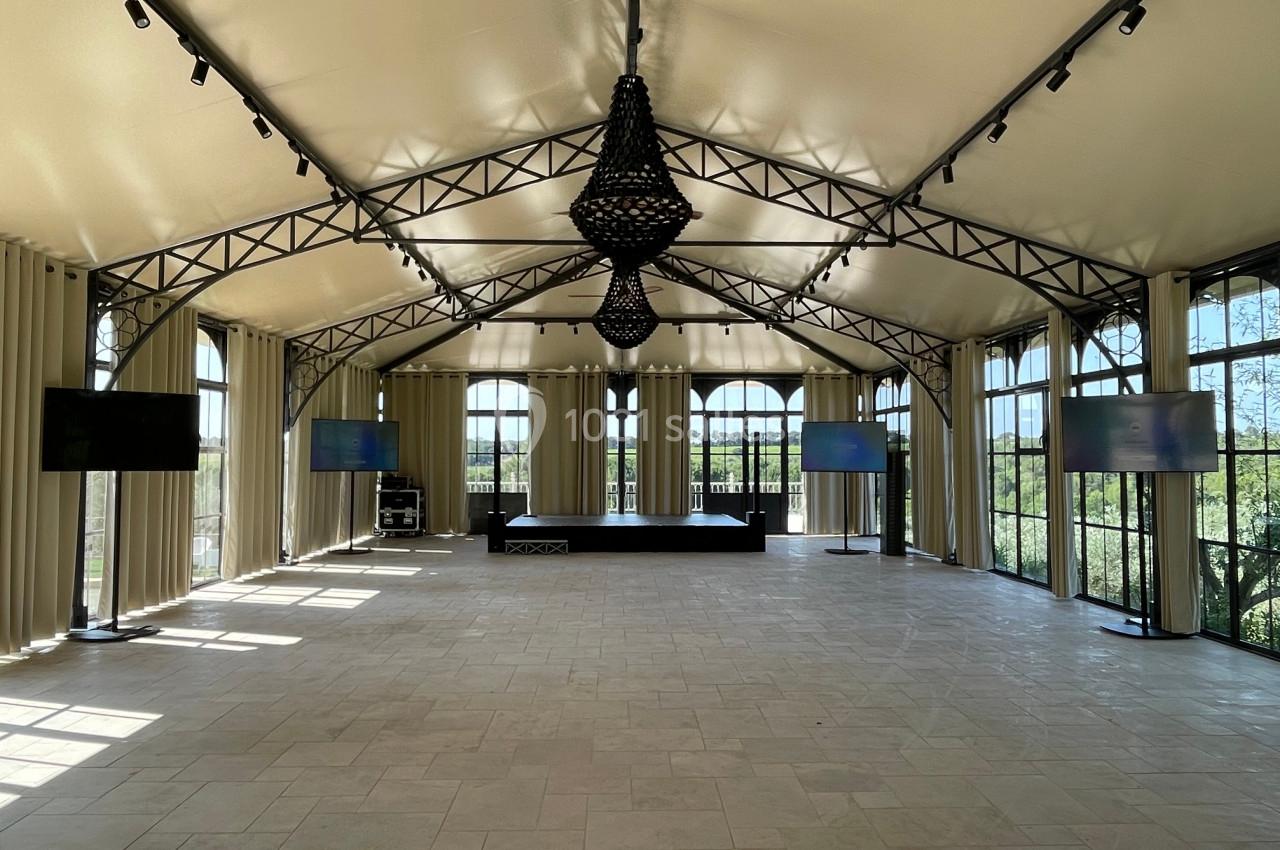 Salle lumineuse avec toit en toile, grandes baies vitrées, scène centrale et écrans disposés sur les côtés.