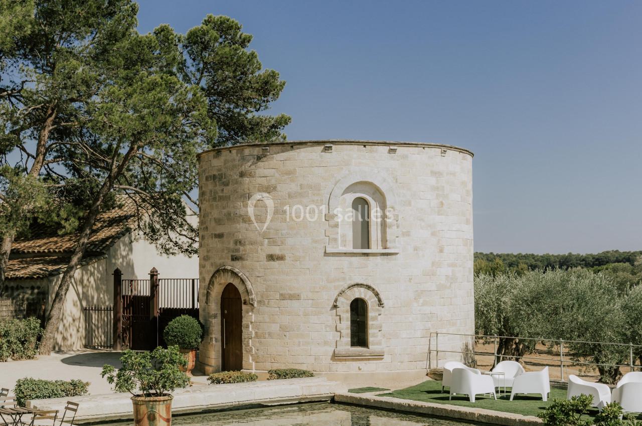 Tour en pierre entourée de végétation, avec bassin d'eau, chaises et tables en métal sur une terrasse ensoleillée.