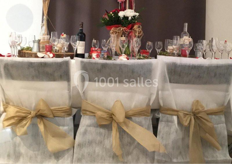 Location salle Rognes (Bouches-du-Rhône) - Domaine Naïs #14 Chaises décorées de nœuds dorés devant une table dressée avec vaisselle, bouteilles et centre de table floral.