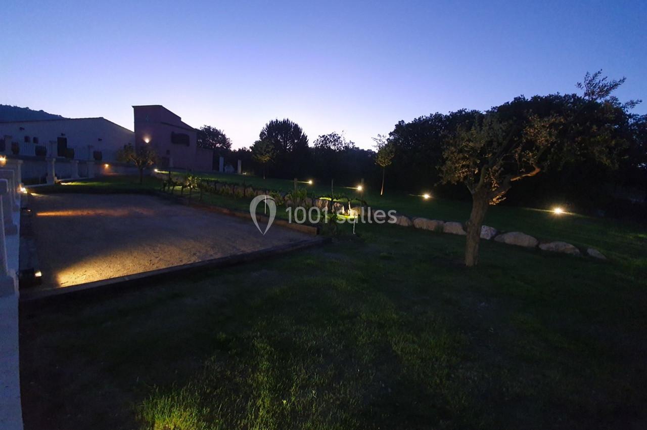 Location salle Rognes (Bouches-du-Rhône) - Domaine Naïs #13 Vue d'un jardin éclairé au crépuscule, avec des arbres, une pelouse, un terrain sablé et des vignes alignées.