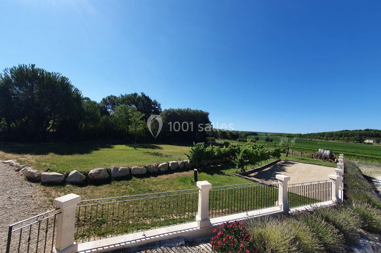 Location salle Rognes (Bouches-du-Rhône) - Domaine Naïs #7 Vue d'un jardin avec pelouse, arbres, allée en gravier, clôture et paysage de campagne sous un ciel bleu.