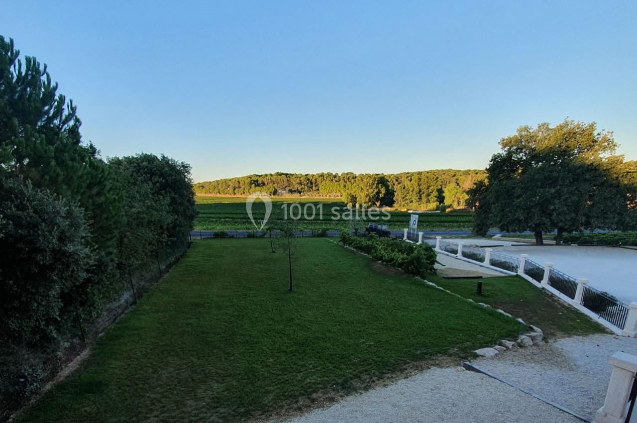 Location salle Rognes (Bouches-du-Rhône) - Domaine Naïs #4 Vue d'un jardin avec pelouse, arbres et allée gravillonnée, donnant sur des vignes et une forêt à l'horizon.
