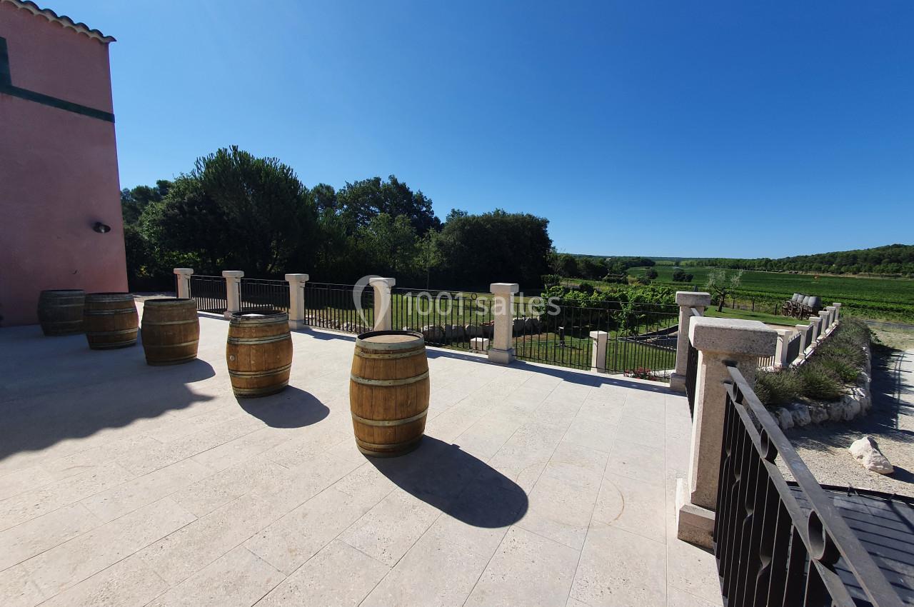 Location salle Rognes (Bouches-du-Rhône) - Domaine Naïs #19 Terrasse en pierre avec des tonneaux en bois, bordée d'une balustrade, donnant sur un jardin et des champs verdoyants.
