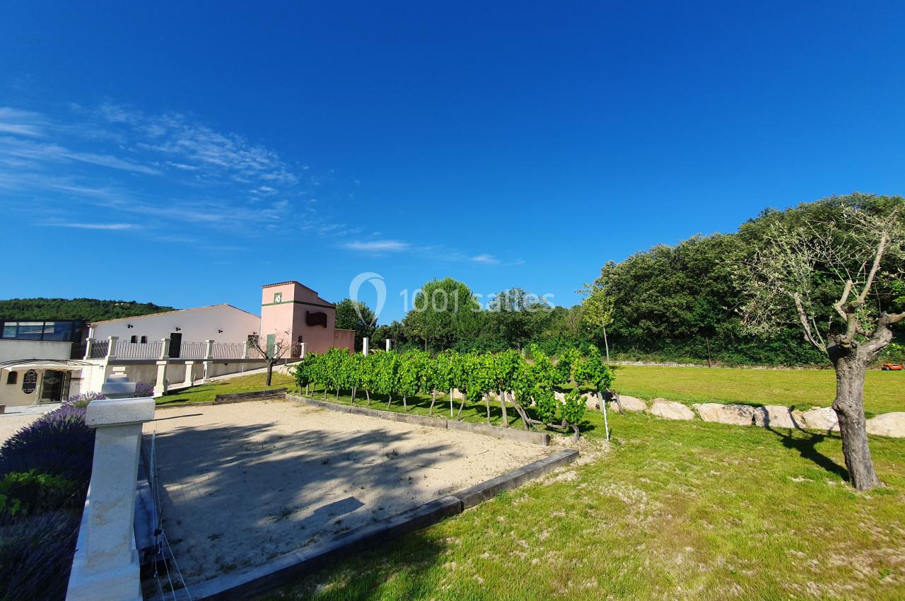 Location salle Rognes (Bouches-du-Rhône) - Domaine Naïs #22 Vue d'un domaine avec bâtiment rose, rangées de vignes, pelouse et ciel bleu dégagé en arrière-plan.