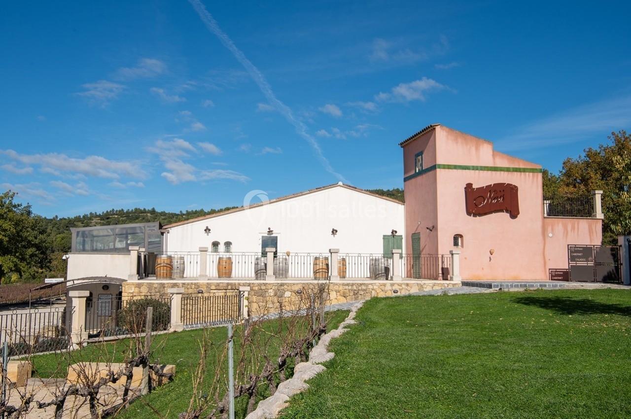 Bâtiment viticole avec façade rose et blanche, entouré de vignes et d'une pelouse sous un ciel bleu.
