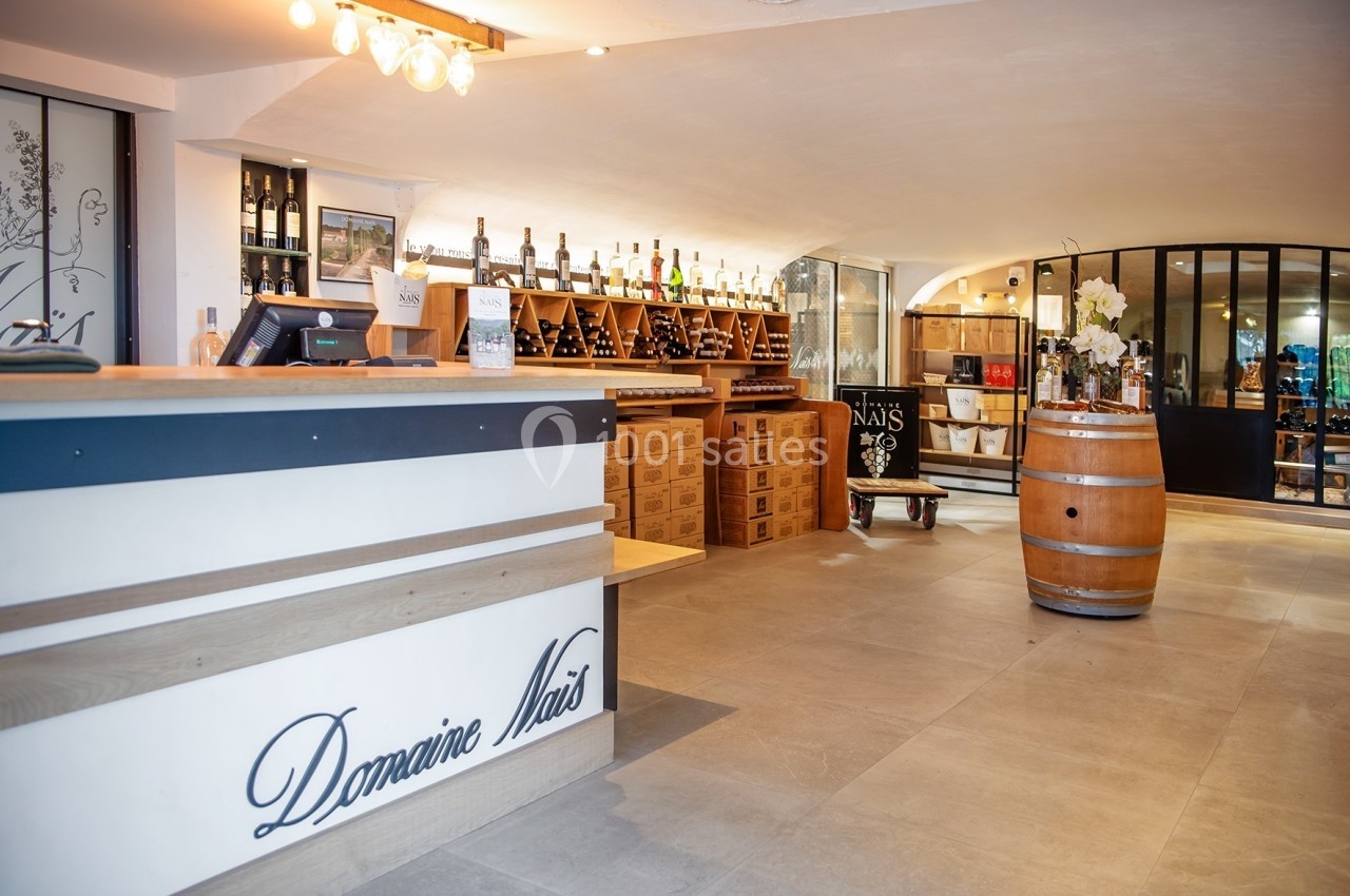 Location salle Rognes (Bouches-du-Rhône) - Domaine Naïs #3 Intérieur d'une cave à vin avec comptoir, étagères de bouteilles, tonneau en bois et décoration lumineuse.