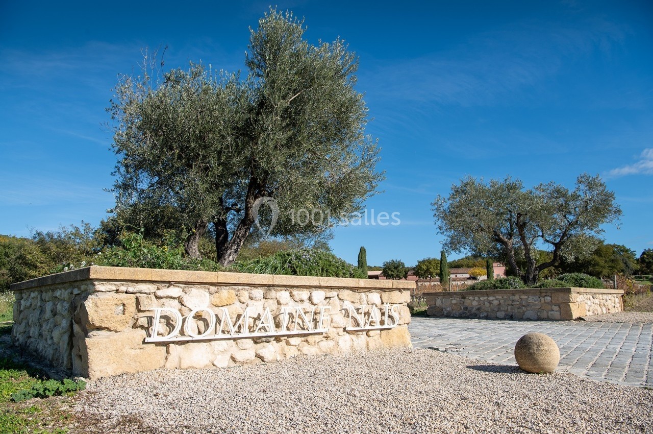 Location salle Rognes (Bouches-du-Rhône) - Domaine Naïs #13 Mur en pierre avec l'inscription ’Domaine’, entouré d'oliviers et d'un chemin pavé sous un ciel bleu.
