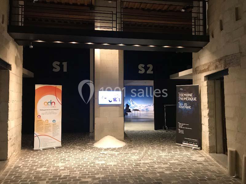 Entrée d'un espace d'exposition avec deux salles, affiches informatives et sol pavé sous un éclairage tamisé.