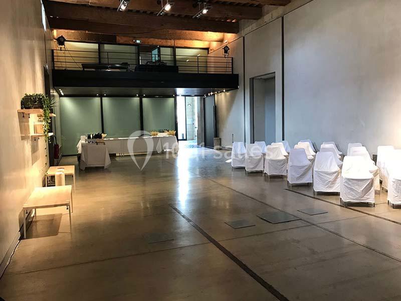 Salle lumineuse avec sol en béton, chaises blanches alignées, tables dressées au fond et mezzanine en bois.